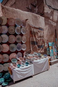 A vibrant street market display featuring an array of intricately designed ceramic plates and bowls arranged on two tables. To the left, numerous plates are mounted on a wall, showcasing various colorful patterns. A woven textile hangs next to them, and to the right, small framed artworks are visible on the ground.