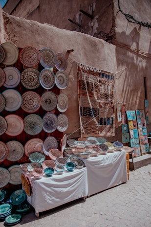 A vibrant street market display featuring an array of intricately designed ceramic plates and bowls arranged on two tables. To the left, numerous plates are mounted on a wall, showcasing various colorful patterns. A woven textile hangs next to them, and to the right, small framed artworks are visible on the ground.