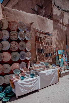 A vibrant street market display featuring an array of intricately designed ceramic plates and bowls arranged on two tables. To the left, numerous plates are mounted on a wall, showcasing various colorful patterns. A woven textile hangs next to them, and to the right, small framed artworks are visible on the ground.
