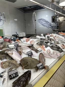 A fish market display with a variety of fresh fish laid out on a bed of ice. Each type of fish has a label with pricing in euros. The surrounding area has white tiled walls decorated with fish illustrations, and industrial lighting brightens the space.
