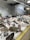 A fish market display with a variety of fresh fish laid out on a bed of ice. Each type of fish has a label with pricing in euros. The surrounding area has white tiled walls decorated with fish illustrations, and industrial lighting brightens the space.