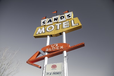 A vintage motel sign for 'Sands Motel' stands against a clear sky. The sign has a prominent yellow background with bold, black lettering, and an additional arrow-shaped orange sign below it. There are small triangular flags on top, adding to its retro aesthetic. A smaller sign at the bottom reads 'AAA Approved' and 'TV & Phones'.