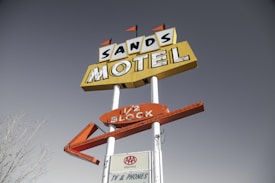 A vintage motel sign for 'Sands Motel' stands against a clear sky. The sign has a prominent yellow background with bold, black lettering, and an additional arrow-shaped orange sign below it. There are small triangular flags on top, adding to its retro aesthetic. A smaller sign at the bottom reads 'AAA Approved' and 'TV & Phones'.