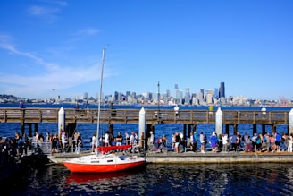 A marina with a red sailboat docked in the foreground. Numerous people are gathered along a pier, socializing and enjoying the sunny weather. In the background, an urban skyline with various skyscrapers and the Space Needle is visible across a large body of water under a clear blue sky.