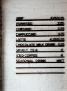 A coffee shop menu displayed on a white brick wall features various drink options and their prices. The menu items include drip coffee, espresso, cortado, cappuccino, latte, chocolate milk drink, spirit tea, iced coffee, and a seasonal drink. Prices are listed next to each beverage.