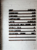 A coffee shop menu displayed on a white brick wall features various drink options and their prices. The menu items include drip coffee, espresso, cortado, cappuccino, latte, chocolate milk drink, spirit tea, iced coffee, and a seasonal drink. Prices are listed next to each beverage.
