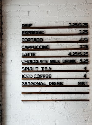 A coffee shop menu displayed on a white brick wall features various drink options and their prices. The menu items include drip coffee, espresso, cortado, cappuccino, latte, chocolate milk drink, spirit tea, iced coffee, and a seasonal drink. Prices are listed next to each beverage.