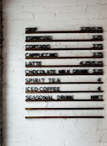 A coffee shop menu displayed on a white brick wall features various drink options and their prices. The menu items include drip coffee, espresso, cortado, cappuccino, latte, chocolate milk drink, spirit tea, iced coffee, and a seasonal drink. Prices are listed next to each beverage.
