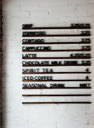 A coffee shop menu displayed on a white brick wall features various drink options and their prices. The menu items include drip coffee, espresso, cortado, cappuccino, latte, chocolate milk drink, spirit tea, iced coffee, and a seasonal drink. Prices are listed next to each beverage.