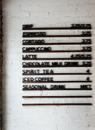 A coffee shop menu displayed on a white brick wall features various drink options and their prices. The menu items include drip coffee, espresso, cortado, cappuccino, latte, chocolate milk drink, spirit tea, iced coffee, and a seasonal drink. Prices are listed next to each beverage.