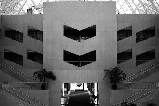 A monochromatic architectural interior with geometric shapes and symmetry. Two staircases on either side lead upwards, flanked by potted palm trees. Large pattern windows are present in the center, giving the design a modern and minimalistic feel. The ceiling features a large, structured grid of glass, allowing natural light to pour in.