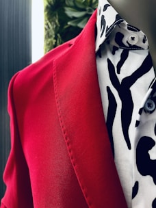 A close-up view of a red blazer paired with a white shirt featuring a bold black pattern. The fabric texture and details are visible, and there is a blurred background with a hint of greenery.