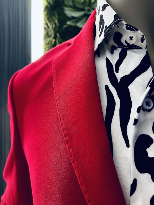 A close-up view of a red blazer paired with a white shirt featuring a bold black pattern. The fabric texture and details are visible, and there is a blurred background with a hint of greenery.