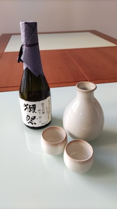 A sake set consisting of a bottle wrapped with a purple paper top, a ceramic carafe, and two small matching cups is placed on a glass table. The background features a wooden table frame.