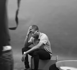 A person is seated on a box, resting their elbow on their knee and their hand on their head, appearing to be deep in thought or contemplation. Glasses adorn their face and they are wearing a watch. The setting has a minimalistic, neutral background with some indistinct objects in the foreground.