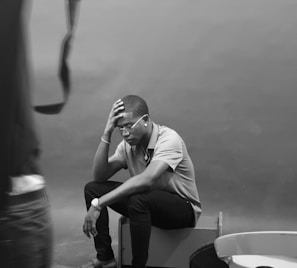 A person is seated on a box, resting their elbow on their knee and their hand on their head, appearing to be deep in thought or contemplation. Glasses adorn their face and they are wearing a watch. The setting has a minimalistic, neutral background with some indistinct objects in the foreground.