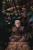A weathered stone statue of an elderly monk is surrounded by dense foliage. The monk is seated in a meditative posture, wearing a traditional robe, with a serene and wise expression. The environment is lush, with various shades of green from the leaves, giving an impression of an overgrown forest or garden.
