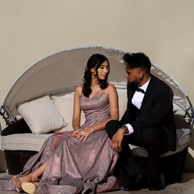 A couple sits on an outdoor wicker sofa with white cushions. The woman is wearing a shimmering pink evening gown and has styled black hair. The man is dressed in a formal black suit with a white shirt and bow tie. They are looking at each other, creating a relaxed and intimate atmosphere. The background is a plain beige wall.