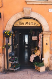 A quaint storefront with an arched entrance displaying 'Zia Mame' above the door. The building facade is a warm orange with stone accents around the arch. Potted flowers in various colors adorn the entrance, and a chalkboard sign indicates flowers for sale at 3.50. Inside, a rustic interior with a welcoming sign can be seen, along with decor items like baskets and cloth.