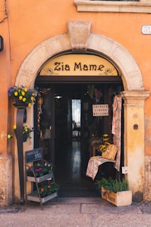 A quaint storefront with an arched entrance displaying 'Zia Mame' above the door. The building facade is a warm orange with stone accents around the arch. Potted flowers in various colors adorn the entrance, and a chalkboard sign indicates flowers for sale at 3.50. Inside, a rustic interior with a welcoming sign can be seen, along with decor items like baskets and cloth.