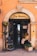 A quaint storefront with an arched entrance displaying 'Zia Mame' above the door. The building facade is a warm orange with stone accents around the arch. Potted flowers in various colors adorn the entrance, and a chalkboard sign indicates flowers for sale at 3.50. Inside, a rustic interior with a welcoming sign can be seen, along with decor items like baskets and cloth.