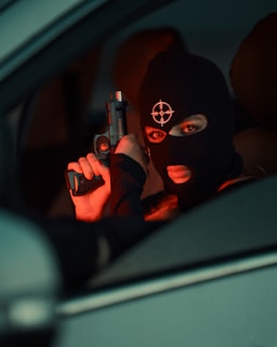 A person wearing a black ski mask with a target symbol holds a handgun inside a vehicle. The lighting is dim with a red tint, suggesting a tense or dangerous atmosphere.