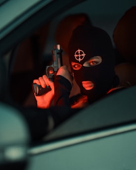 A person wearing a black ski mask with a target symbol holds a handgun inside a vehicle. The lighting is dim with a red tint, suggesting a tense or dangerous atmosphere.
