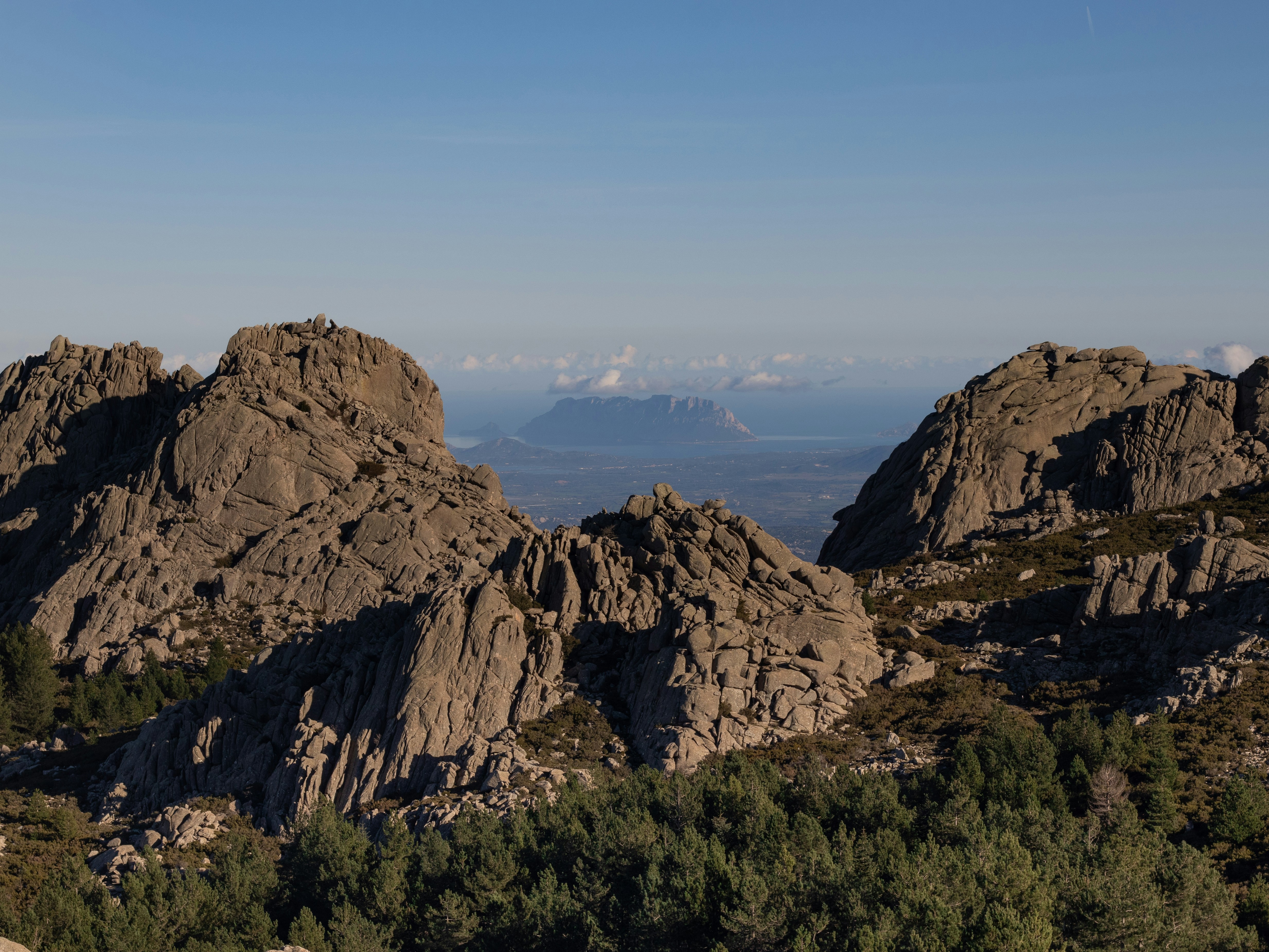 a mountain range with trees and mountains in the background, Tavolara island and Olbia harbor seen from behind the granite rock formations at the top of mount Limbara in Sardinia.