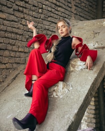 A person poses against a textured brick wall, sitting on a sloped surface. They wear a striking outfit featuring a black top with oversized red ruffled sleeves and red wide-leg pants. The pose is casual yet stylish, with one arm raised and the other relaxed. The setting appears industrial with a modern fashion aesthetic.