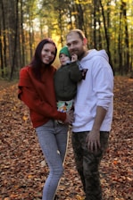 A family of three is standing on a forest floor covered with autumn leaves. The woman on the left is wearing a red sweater and jeans, smiling, while the man on the right in a white hoodie has one arm around a child dressed in a green jacket and beanie.