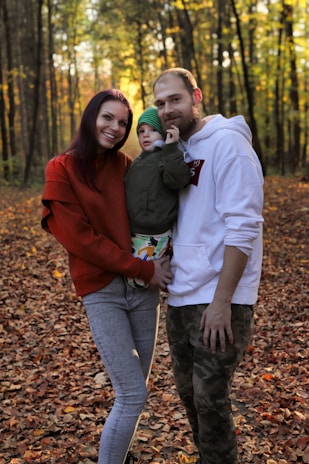 A family of three is standing on a forest floor covered with autumn leaves. The woman on the left is wearing a red sweater and jeans, smiling, while the man on the right in a white hoodie has one arm around a child dressed in a green jacket and beanie.