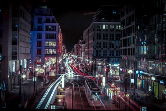 A bustling urban street at night with streaks of light from moving vehicles creating a lively atmosphere. Tall buildings flank both sides of the street, illuminated with colorful lights and signage. The scene is vibrant, capturing the dynamic energy of city life.