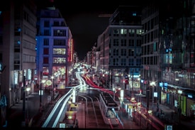 A bustling urban street at night with streaks of light from moving vehicles creating a lively atmosphere. Tall buildings flank both sides of the street, illuminated with colorful lights and signage. The scene is vibrant, capturing the dynamic energy of city life.