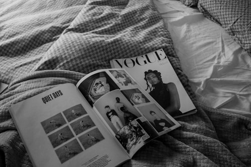 A black and white photograph features an open magazine resting on a bed with textured blankets. The magazine pages display a title reading 'QUIET DAYS' alongside various images, and the opposite page shows a cover with a portrait photograph. The setting appears casual and relaxed.