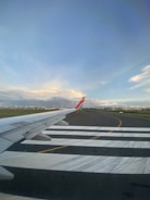 An airplane's wing is visible in the foreground as it taxis on a runway. The sky is mostly clear with soft clouds, and there are airport buildings and other stationary planes in the distance.