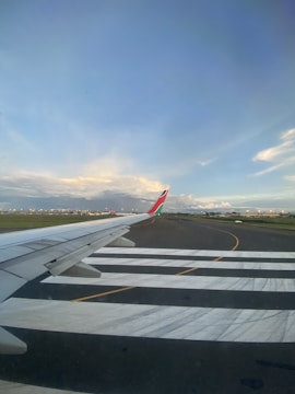 An airplane's wing is visible in the foreground as it taxis on a runway. The sky is mostly clear with soft clouds, and there are airport buildings and other stationary planes in the distance.
