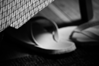 A pair of flip-flops placed on the floor next to a piece of upholstered furniture with a textured, grid-patterned fabric. The image is in black and white, creating a high-contrast effect with soft focus on the edges.