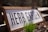 A wooden sign with the words 'HERB GARDEN' is positioned at an angle, partially buried in soil. In front of the sign, green grass or herb shoots emerge from the soil, with sunlight casting shadows behind them. The background is blurred, suggesting a garden environment.