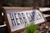 A wooden sign with the words 'HERB GARDEN' is positioned at an angle, partially buried in soil. In front of the sign, green grass or herb shoots emerge from the soil, with sunlight casting shadows behind them. The background is blurred, suggesting a garden environment.
