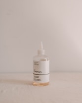 A bottle of The Ordinary brand skincare product with minimalist design, featuring a white label with black text. The bottle is transparent with a small amount of light orange liquid inside, set against a plain, softly lit backdrop.