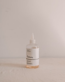 A bottle of The Ordinary brand skincare product with minimalist design, featuring a white label with black text. The bottle is transparent with a small amount of light orange liquid inside, set against a plain, softly lit backdrop.