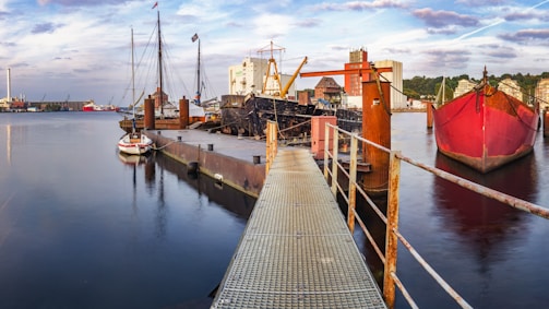 A waterfront scene with several boats docked at a pier. A red cargo ship is prominent on the right side, while a smaller sailboat is visible closer to the pier. The backdrop features industrial buildings and cranes, with more ships in the distance. The water reflects the sky, which is partly cloudy with hints of blue.