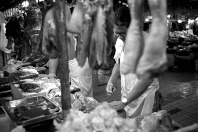 A black and white photograph of a butcher working in a market. Various cuts of meat hang from hooks and are displayed on a table. The butcher appears focused on preparing the meat, and the image captures the bustling environment of the market with other stands visible in the background.