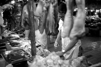 A black and white photograph of a butcher working in a market. Various cuts of meat hang from hooks and are displayed on a table. The butcher appears focused on preparing the meat, and the image captures the bustling environment of the market with other stands visible in the background.