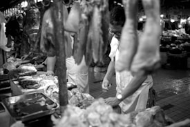 A black and white photograph of a butcher working in a market. Various cuts of meat hang from hooks and are displayed on a table. The butcher appears focused on preparing the meat, and the image captures the bustling environment of the market with other stands visible in the background.