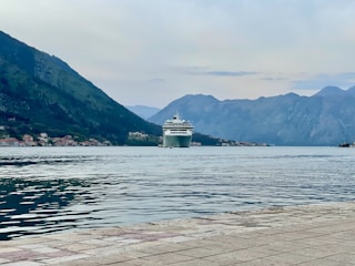 A large cruise ship is positioned in a calm bay surrounded by towering mountains. Small buildings are visible along the shoreline, and the foreground features a stone or concrete waterfront.