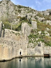 A sturdy, historical stone fortress is built into a rugged mountainside. The fortress features old, weathered masonry, arched doorways, and is surrounded by green vegetation. The reflection of part of the structure can be seen in the calm water below. The sky above is clear with a hint of light clouds.