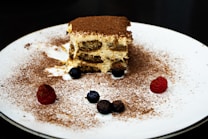 A plate with a piece of tiramisu, covered with a dusting of cocoa. The dessert is surrounded by a few raspberries and blueberries.