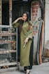 A woman with long dark hair stands next to a display case filled with watches, wearing an olive green &aacute;o d&agrave;i and holding a bouquet of red flowers. The setting appears to be a sidewalk shop with various signs and exposed cables in the background.