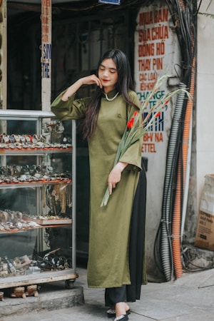 A woman with long dark hair stands next to a display case filled with watches, wearing an olive green áo dài and holding a bouquet of red flowers. The setting appears to be a sidewalk shop with various signs and exposed cables in the background.